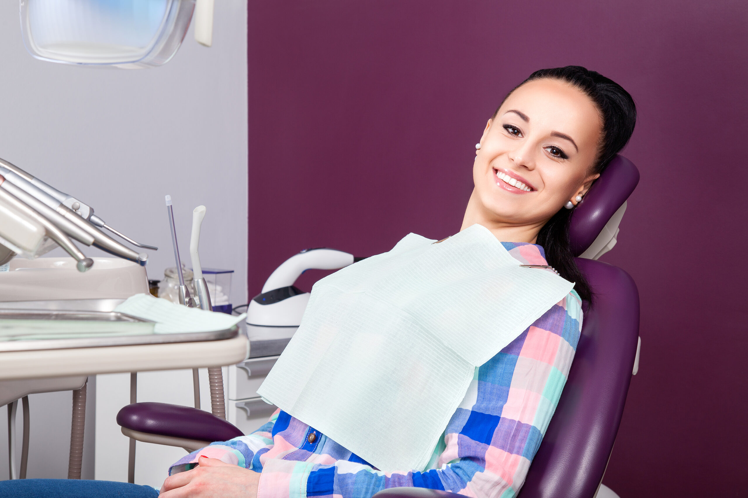 Young woman patient in checkered shirt with perfect straight white teeth waiting for dentist in dental chair and smiling relaxed ready for a check-up. Beautiful woman smile
